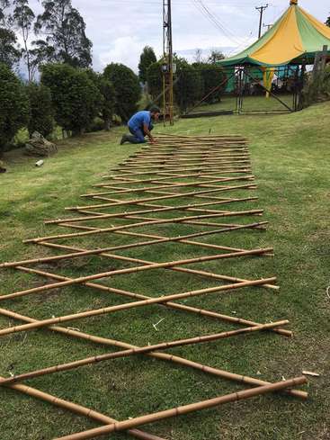 A man is kneeling on the grass, assembling a large bamboo lattice structure, with a yellow and green tent in the background on a cloudy day.