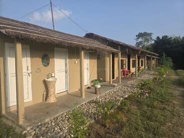 This image shows a row of rustic, thatched-roof cottages with white doors, outdoor sinks, and a pebble-lined walkway, surrounded by greenery, under a clear sky.