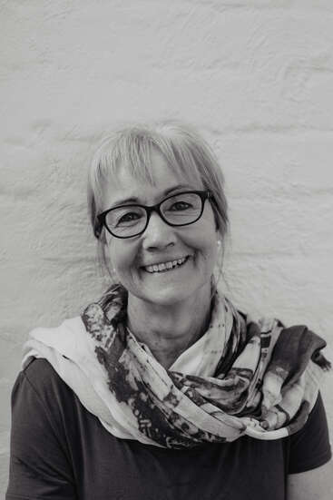 The image is a black-and-white portrait of a woman with short hair, wearing glasses and a scarf, standing in front of a white wall.