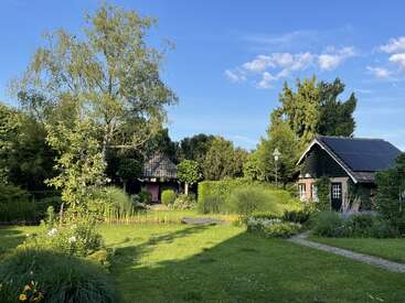 Eine bezaubernde ländliche Szene mit einem kleinen Holzhaus, einem üppigen grünen Garten, blühenden Blumen, hohen Bäumen, einem klaren blauen Himmel und einer ruhigen, sonnigen Atmosphäre.