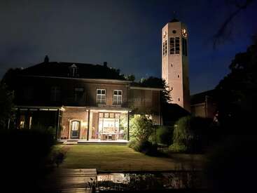 Ein großes, nachts warm beleuchtetes Backsteinhaus mit einem hohen Uhrenturm daneben, umgeben von einem Garten und Bäumen, alles unter einem dunklen Himmel.