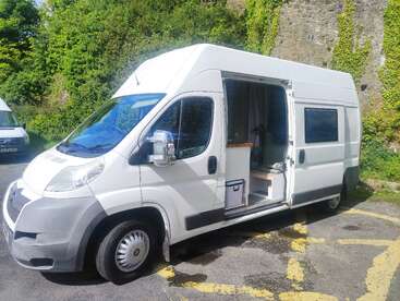Un camping-car blanc est garé dans un endroit ensoleillé. Sa porte latérale est ouverte, révélant un intérieur confortable et compact. En arrière-plan, de la verdure et un mur de pierre.