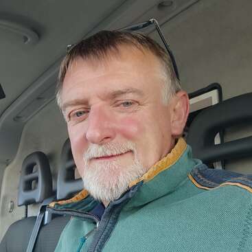 A middle-aged man with short hair and a trimmed white beard smiles inside a vehicle. He wears a green jacket and glasses rest on his head.