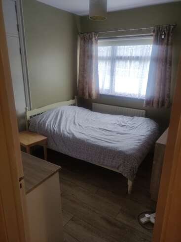 Une petite chambre à coucher avec un lit simple, une literie légère, des planchers en bois, des murs verts, des rideaux à fleurs, une table de nuit en bois, un radiateur et une fenêtre avec des rideaux en dentelle.