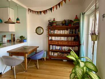 Dieser gemütliche Raum verfügt über ein Bücherregal aus Holz voller Bücher, einen rustikalen Tisch mit zwei Stühlen, Hängepflanzen, festliche Girlanden und farbenfrohe Deko-Akzente. Eine einladende Atmosphäre.