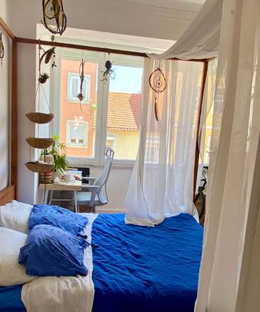 Ein gemütliches Schlafzimmer mit einem Himmelbett, blauer Bettwäsche, weißen Vorhängen, Traumfängern, hängenden Körben, einem Schreibtisch, Pflanzen und einem großen Fenster, durch das Sonnenlicht hereinscheint.