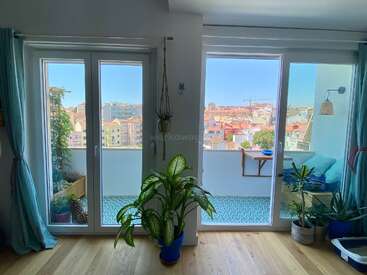 Helle Wohnung mit großen Glastüren, die auf einen Balkon führen, gemütliche Sitzgelegenheiten und Topfpflanzen. Blick auf die Stadt, türkisfarbene Vorhänge, viel Tageslicht und dekorative blaue Fliesen.
