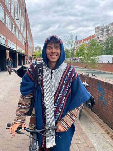 Un joven sonriente con un poncho de colores y capucha está de pie con una bicicleta en una calle de la ciudad, con edificios, árboles y un cielo nublado detrás.