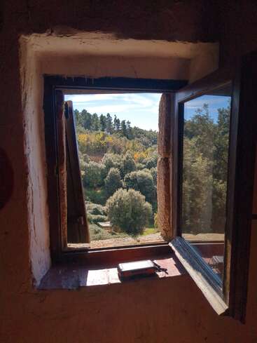 A luz do sol entra por uma janela rústica aberta, iluminando um dispositivo no peitoril. Árvores verdes e exuberantes e um céu azul emolduram uma cena campestre tranquila.