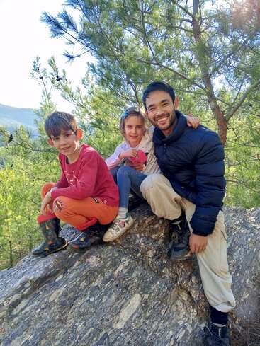 Três pessoas, duas crianças e um adulto, sentam-se em uma grande rocha ao ar livre, cercada por árvores. Elas estão sorrindo, desfrutando juntas de um dia ensolarado na natureza.