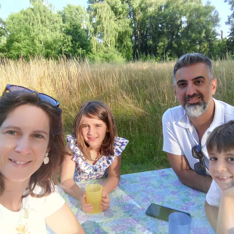 Uma família de quatro pessoas desfruta de um piquenique ensolarado ao ar livre. Eles sorriem para a câmera, sentados em uma mesa cercada por grama alta e árvores verdes exuberantes.