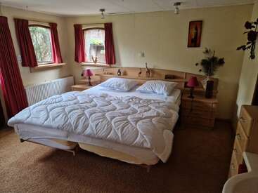 This cozy bedroom features a large double bed with a white comforter, red curtains, wooden furniture, bedside lamps, decorative art, and soft brown carpeting.