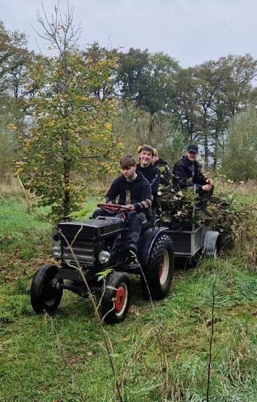 Three young people ride a small tractor pulling a trailer full of branches through a grassy, wooded area. Two are smiling, enjoying their outdoor work.