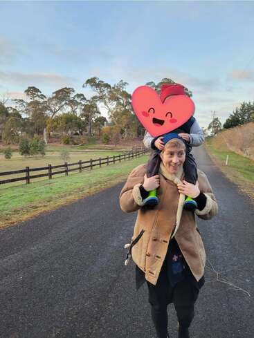 Eine Person geht eine Landstraße entlang und trägt ein Kind auf den Schultern. Das Gesicht des Kindes ist mit einem Herz-Emoji bedeckt. Der Hintergrund ist von Bäumen gesäumt.