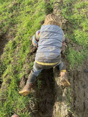 Ein kleines Kind, das einen bunten Pullover, eine blaue Weste und schlammige Stiefel trägt, krabbelt auf Händen und Knien durch ein grasbewachsenes, schlammiges Feld und erkundet mit Begeisterung die freie Natur.