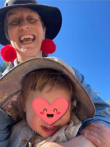 Ein lächelnder Erwachsener mit schwarzem Hut und roten Bommel-Ohrringen posiert im Freien mit einem Kind, dessen Gesicht von einem Herz-Emoji verdeckt wird, unter einem klaren blauen Himmel.