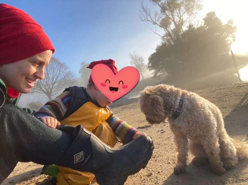 Eine lächelnde Frau mit einem roten Hut sitzt im Freien mit einem Kind, dessen Gesicht von einem Herzaufkleber verdeckt ist, und ein kleiner Hund schaut zu.