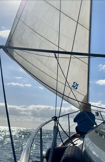 A person in a knitted hat sits at the bow of a sailboat, under bright sunlight. The sail is full, ocean sparkling beneath a blue sky.