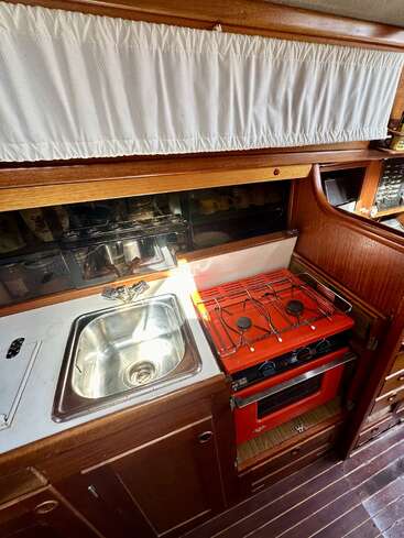This image shows a compact boat galley with a small stainless steel sink, a distinctive red stove-oven unit, wooden cabinetry, and white curtain above. Cozy atmosphere.
