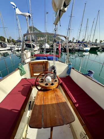 A sailboat cockpit with a wooden table and maroon cushions, docked in a marina, surrounded by other boats under a clear blue sky.