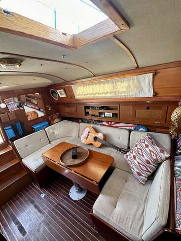This cozy boat interior features a cushioned L-shaped bench, wooden table, decorative pillow, acoustic guitar, books, and sunlight streaming through an open ceiling hatch.