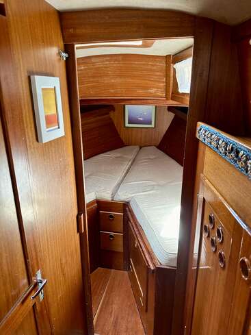 This image shows a cozy wooden boat cabin with a V-shaped bed, storage drawers, wall art, and a small window with a curtain, radiating warmth and comfort.