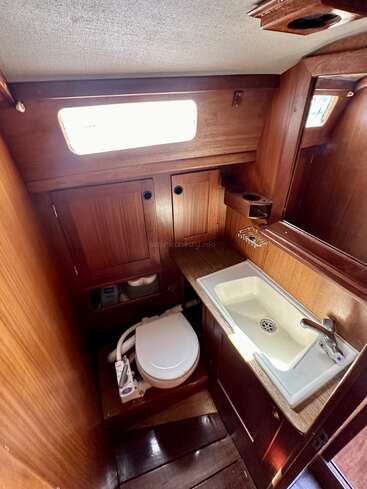 This cozy boat bathroom features wooden paneling, a manual marine toilet, a small rectangular sink with faucet, mirrored cabinet, storage, and a window allowing natural light.