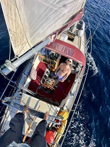 A man stands smiling on a sailboat in the ocean. The photo is taken from above, showing another person’s feet on the mast, blue water, and sails.