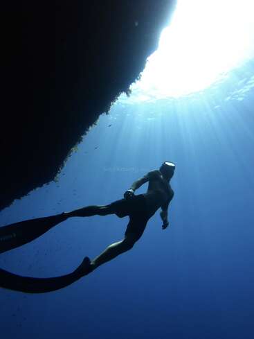 A snorkeler glides underwater near a rocky wall, illuminated by sunlight streaming through the ocean’s surface, surrounded by deep blue water and tiny fish.