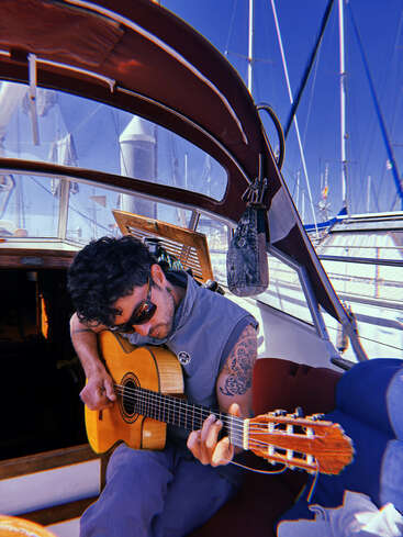 A man with sunglasses and a tattooed arm plays an acoustic guitar on a boat. Sunlight streams in, sailboats and a clear blue sky in the background.