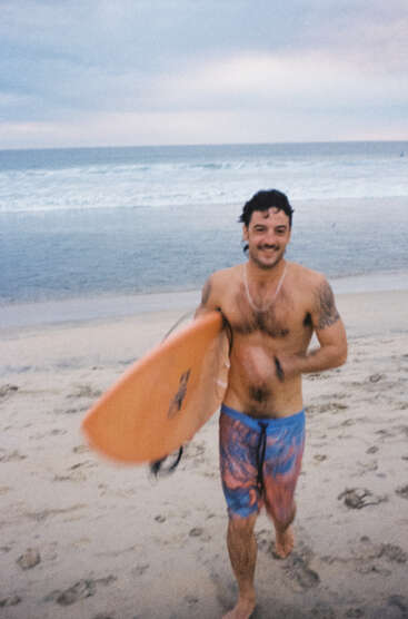 A smiling man with a mustache runs on the sandy beach, holding an orange surfboard, wearing colorful swim trunks. Ocean waves and cloudy sky behind him.