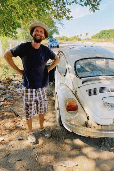 A smiling man in a hat stands beside a weathered vintage Volkswagen Beetle on a sunlit roadside, wearing plaid shorts, a black shirt, and crocs.