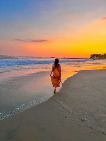 A woman in a flowing orange dress walks barefoot along a serene beach at sunset, with colorful skies reflected on gentle waves and tranquil shoreline.