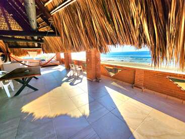 A sunlit beachfront terrace with a thatched roof, brick columns, hammocks, white plastic chairs, and a large wooden table overlooks a sparkling blue ocean.