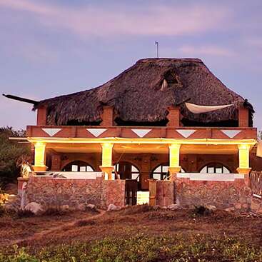 A rustic house with a thatched roof, stone and brick walls, warmly lit columns, sits on a grassy landscape under a soft, evening sky.