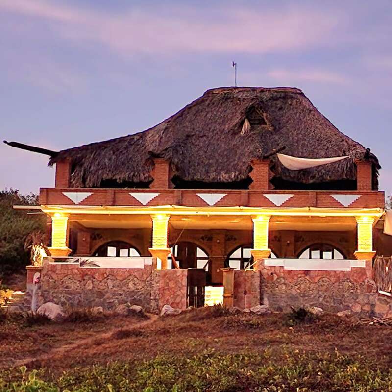 A rustic house with a thatched roof, stone and brick walls, warmly lit columns, sits on a grassy landscape under a soft, evening sky.