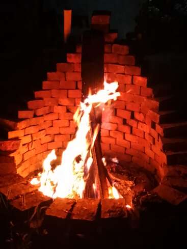 À noite, uma fogueira brilhante arde dentro de uma fogueira circular de tijolos. Os troncos estão empilhados na vertical, rodeados por chamas quentes e alaranjadas, criando um ambiente aconchegante.