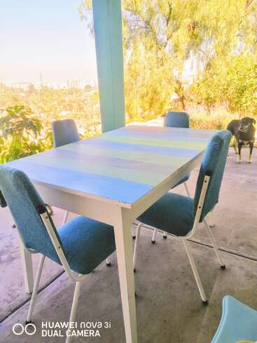 Uma mesa em tons pastéis com quatro cadeiras azuis fica em um pátio à sombra. Lá fora, dá pra ver árvores verdes e a paisagem urbana. Um cachorro preto está parado ali perto, observando.