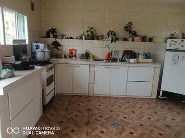 Essa cozinha tem armários brancos, piso de azulejo, geladeira, fogão, plantas nas prateleiras, louças perto da pia, vários utensílios e luz natural.