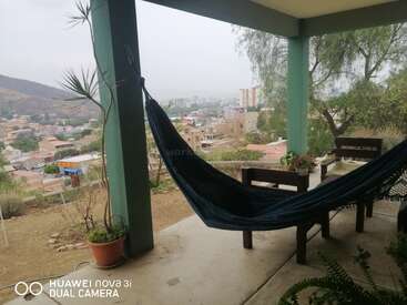 Um pátio coberto aconchegante conta com uma rede escura, cadeiras de madeira, vasos de plantas e uma vista panorâmica para um bairro na encosta e para a paisagem urbana ao longe.