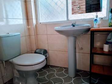 Este banheiro tem um vaso sanitário, um lavatório com pedestal, um cesto de lixo pequeno, um dispensador de sabonete líquido e uma prateleira com produtos de limpeza. A janela deixa a luz natural entrar.