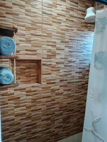 A imagem mostra um banheiro com azulejos marrons estampados, toalhas azuis enroladas em prateleiras rústicas de madeira, um chuveiro branco e uma cortina de chuveiro com desenhos de conchas.