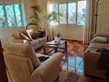 Esta aconchegante sala de estar tem sofás bege, poltronas, uma mesa de centro de madeira, plantas de interior e grandes janelas ensolaradas com cortinas brancas, criando um ambiente acolhedor e convidativo.