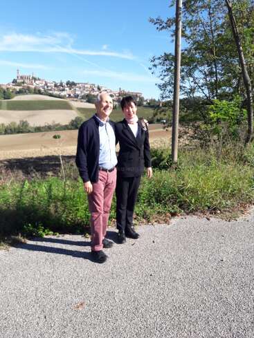 A smiling couple stands on a rural road, surrounded by greenery. In the background, a picturesque hilltop village is visible under a bright blue sky.