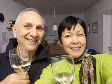 A smiling couple stands together indoors, holding glasses of white wine. They appear happy and relaxed, perhaps celebrating or enjoying a special evening at home.