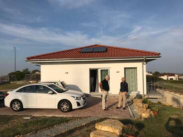 Two men stand outside a modern, single-story house with a red roof and solar panel. A white car is parked in the driveway. Sunny day.