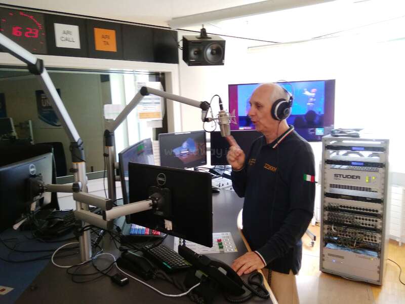 A man wearing headphones speaks into a microphone in a professional radio studio. Multiple monitors, audio equipment, and soundproofed surroundings create a high-tech broadcasting environment.