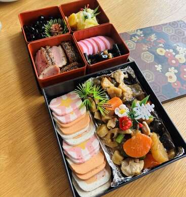 Essa imagem mostra uma refeição tradicional japonesa de Ano Novo, o Osechi Ryori, lindamente disposta em caixas lacadas, com legumes coloridos, carnes e enfeites decorativos.