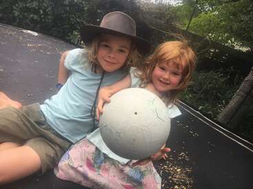 Two children sit on a trampoline outdoors, smiling at the camera. One wears a wide-brimmed hat, while the other holds a large gray ball. Trees surround them.