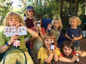 A group of seven children sit outdoors, smiling and holding bottles with labels. One child holds a handwritten sign with Chinese characters. Lush greenery surrounds them.